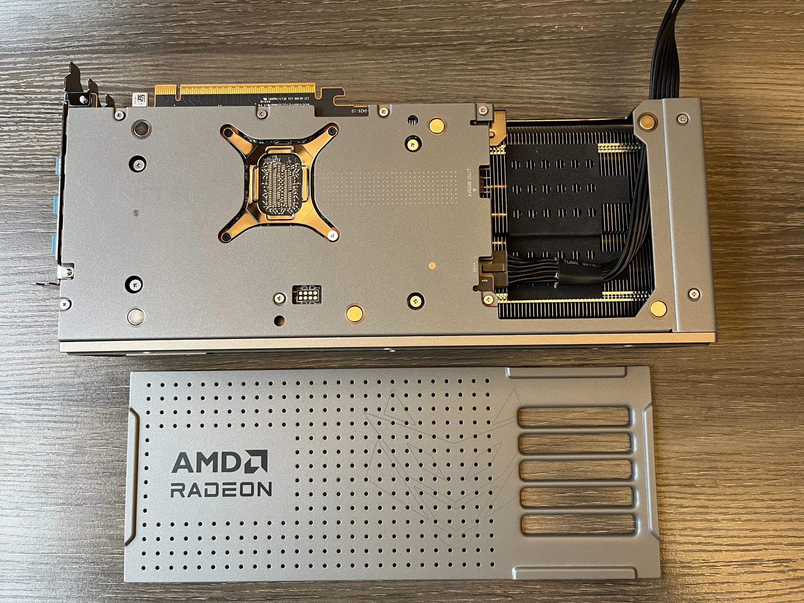 A picture of the Sapphire Nitro+ 9070 XT laying fan-side down on a wooden table. The cosmetic backplate has been removed and is sitting below the card, right-side up. On the right side of the card is a cutout in the structural metal backplate, thourhg which the finstack can be seen 1/2 inch below the plans of the backplate. On the fins are sections of of felt or foam with small holes in them. On the leftside of the cutout there is a 12VHPWR cable pluggd into a power connector, above which is an RGB connector, with nothing plugged into it. The cable of the 12V HPWR connector is folded over on itself, running across the front of the cutout to the right side of the cutout, at which point the fold occurs and it runs stright up and out of the card at the top.