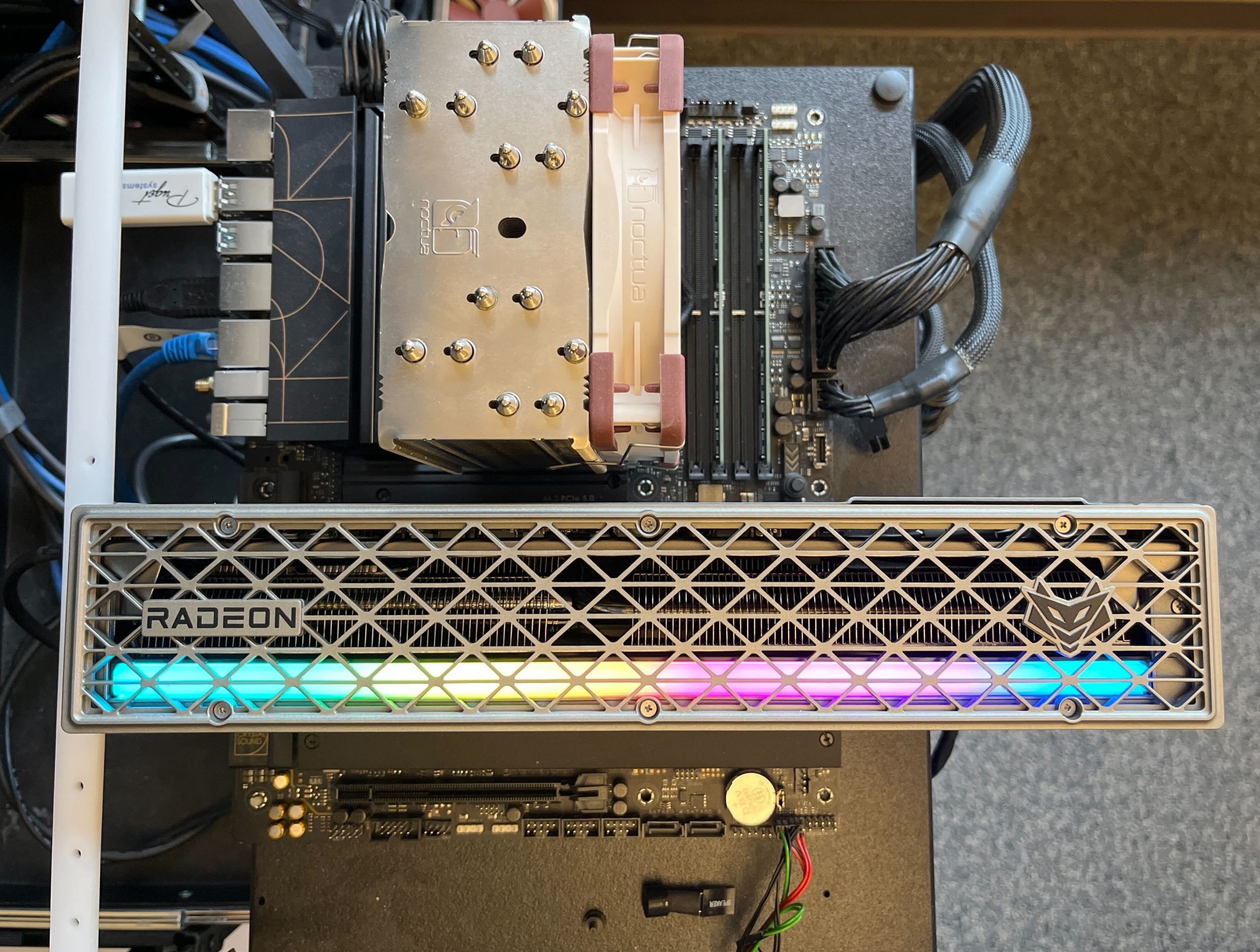 A picture of the test bed: the Sapphire Nitro+ 9070 XT plugged installed in an ASUS ProArt X670 Motherboard, in the top PCIe slot. Above it, a Noctua NH-U12A cooler is visible, with one fan installed on the right-hand side, to the right of which are tow RAM sticks installed in the far right and second-from-the-left slots. There are cables sticking out from the I/O ports on the left side of the motherboard, but you canot see the GPU power cable. The LED light bar on the GPU is well diffused and you cannot see any of the individual LEDs.